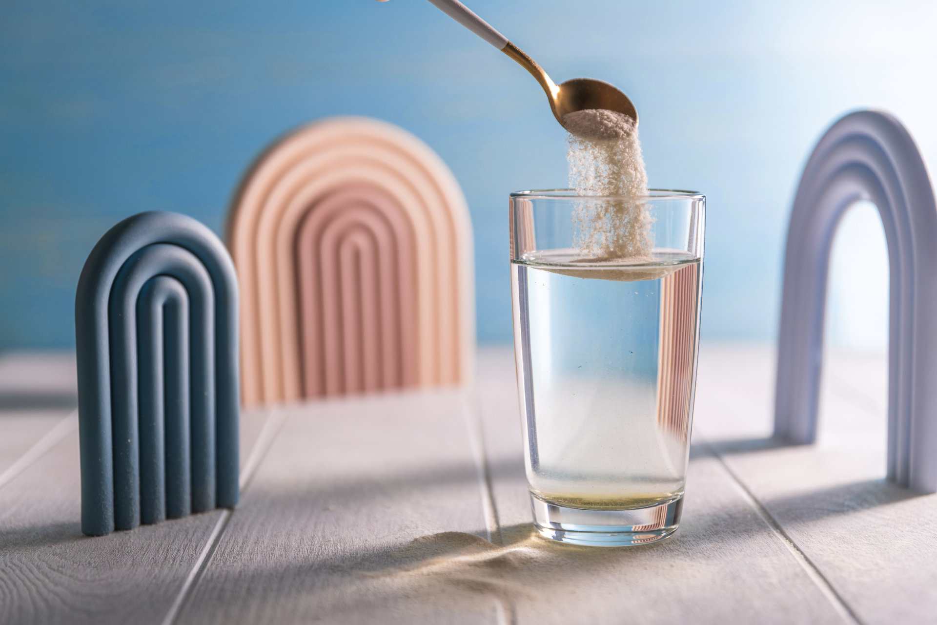A spoon pours ORS powder into a glass of water, displayed with minimalist arch decorations against a light blue background