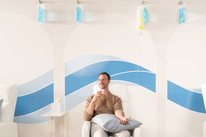 A man sits in a comfortable clinic chair receiving an IV therapy session, holding a drink, and smiling