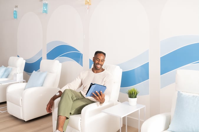 Man relaxing in a bright IV therapy lounge while looking at a tablet, with IV bags hanging overhead and white recliner chairs in the background