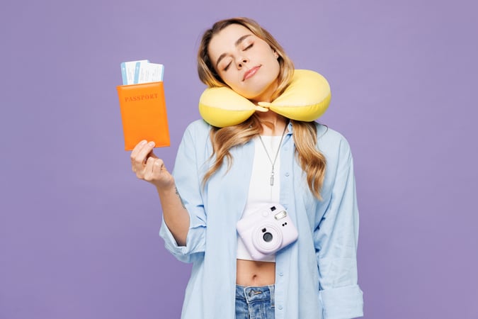 A young woman with her eyes closed wearing a yellow travel neck pillow, holding up an orange passport holder with boarding passes against a solid purple background