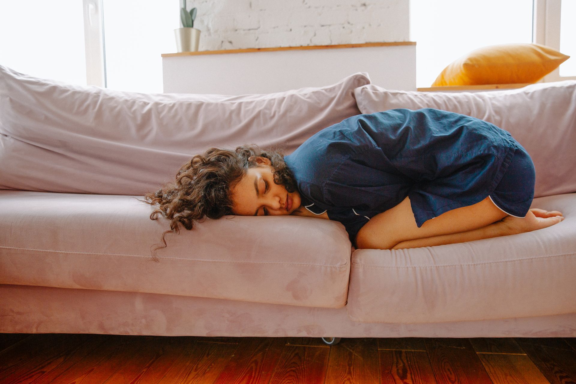 A woman rests on a sofa in pajamas, curled up in menstrual discomfort during the day
