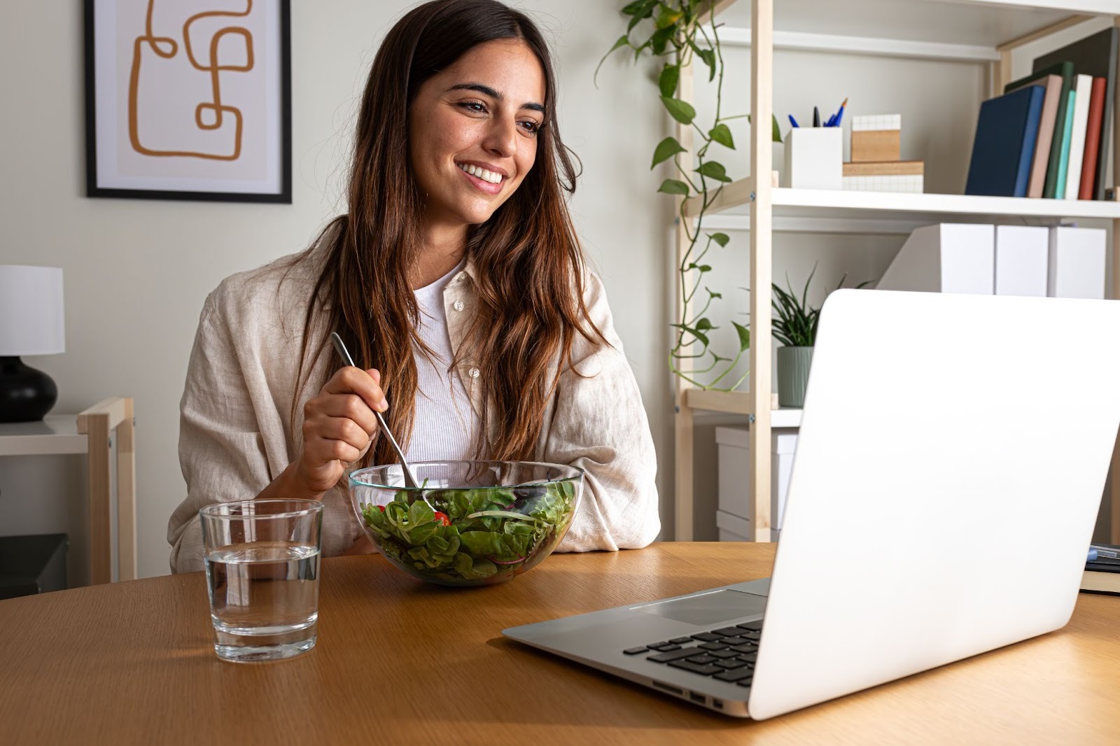 A woman at her desk smiling, eating a salad while on a work call on her laptop.