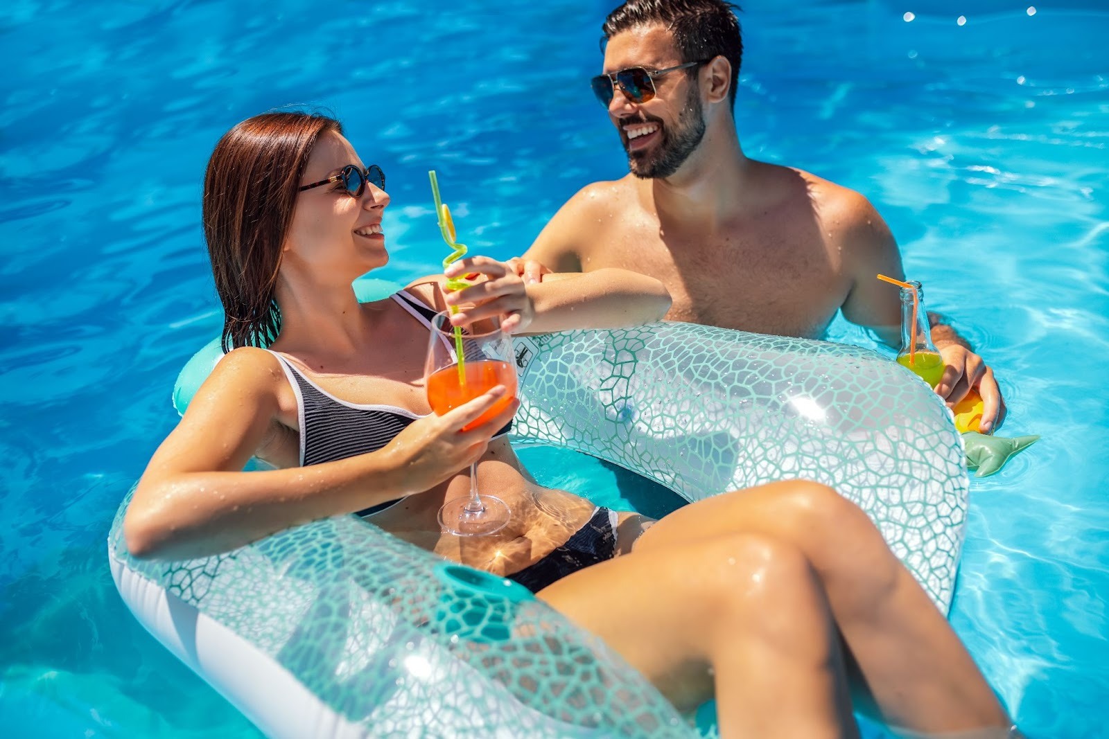 A young couple lounge in a pool, drinks in hand.
