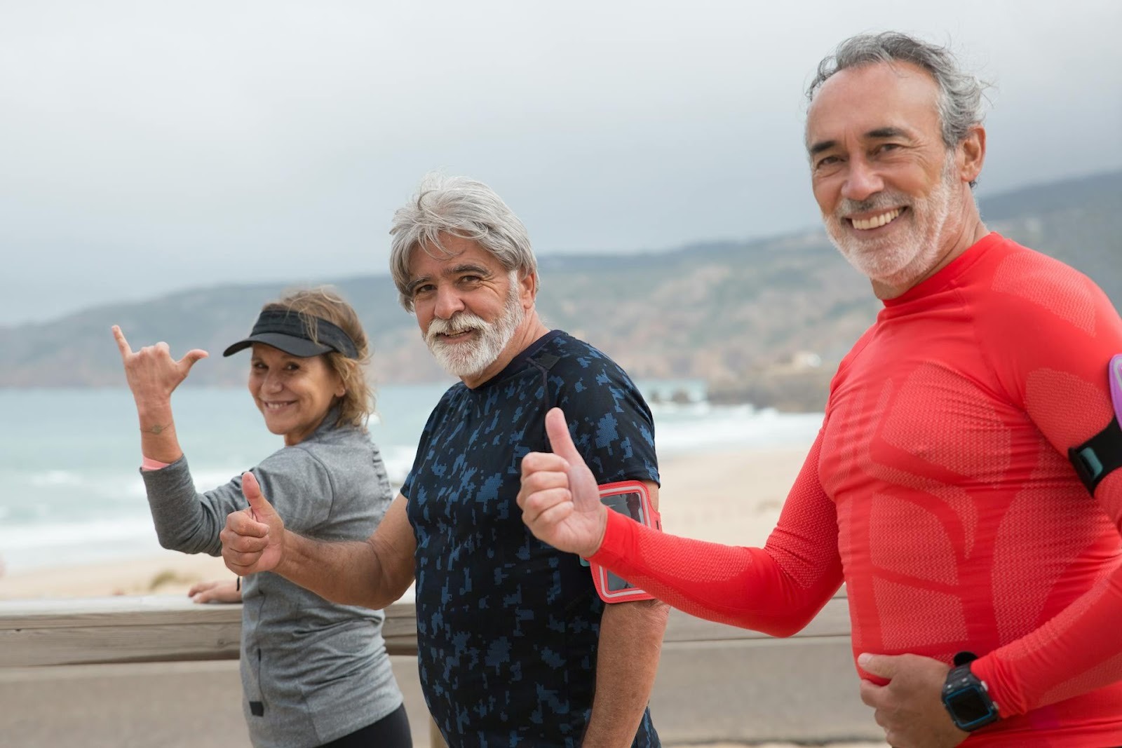 Three older adults in athletic wear, smiling and giving thumbs up while walking or jogging on a beach boardwalk