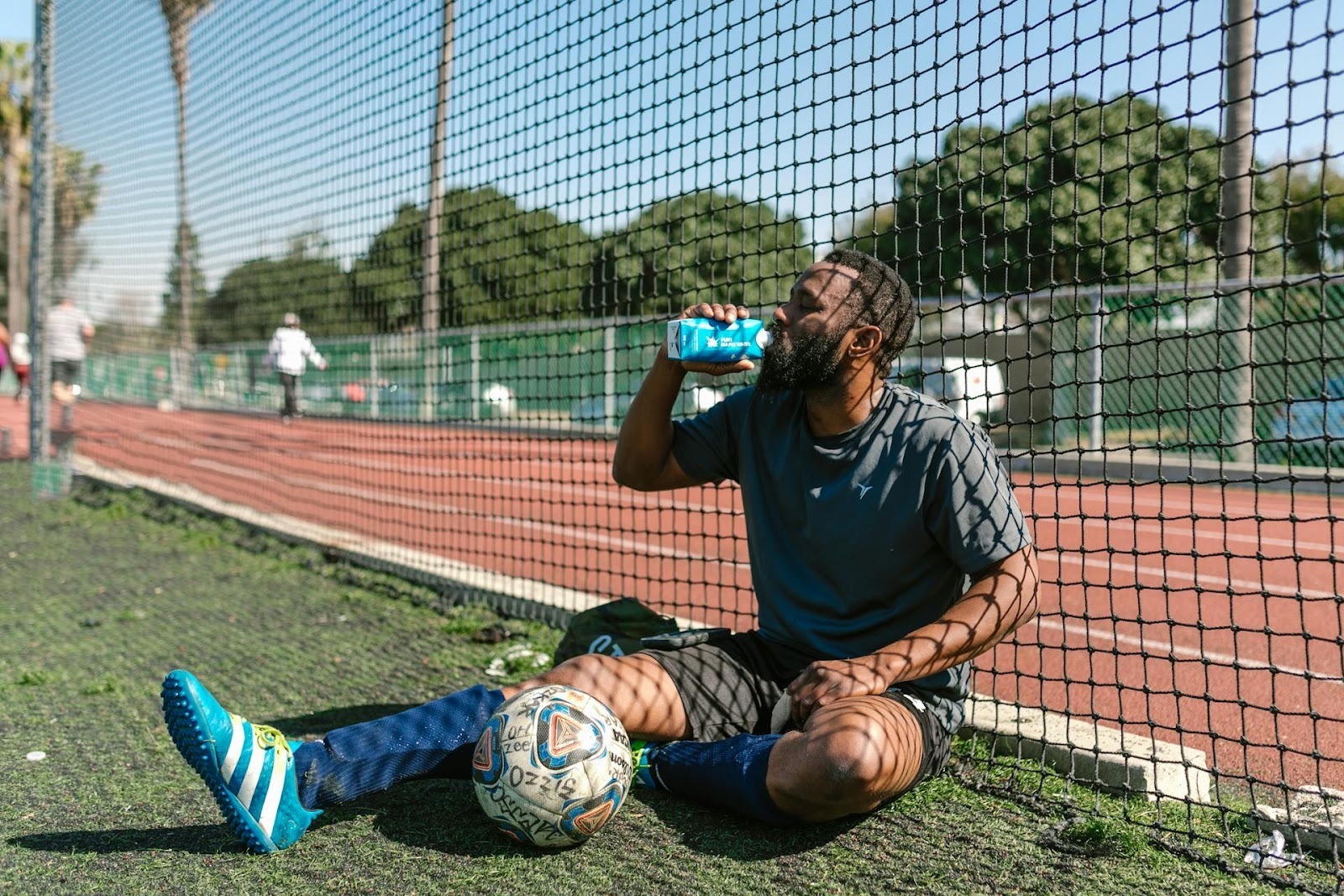 A man in athletic wear sits on artificial turf beside a fenced soccer field, drinking from a blue carton