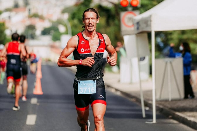 A smiling male athlete competing in a marathon, wearing a black and red racing suit, running on a paved road