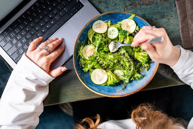 An overhead view of a person eating a fresh green salad from a blue bowl while working on a silver laptop