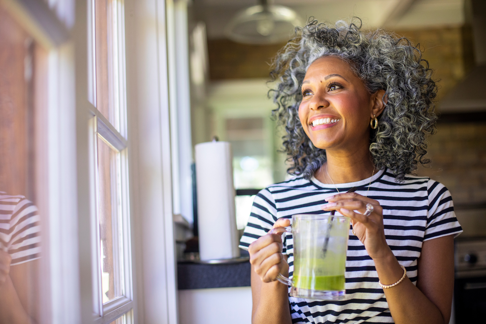 A middle-aged woman with a healthy green smoothie, smiling while looking outside her home