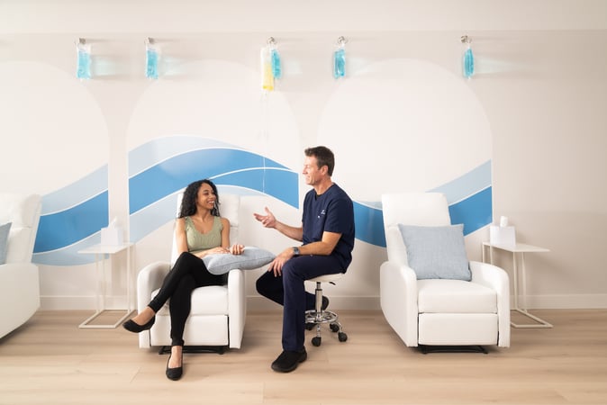 Young smiling woman speaking with a clinician in a modern IV therapy clinic with IV bags hanging behind them.