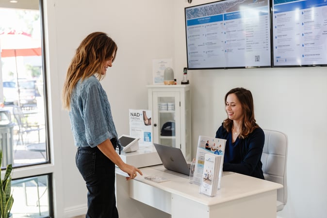 A woman standing at a reception desk speaks with a seated receptionist using a laptop in a bright wellness clinic