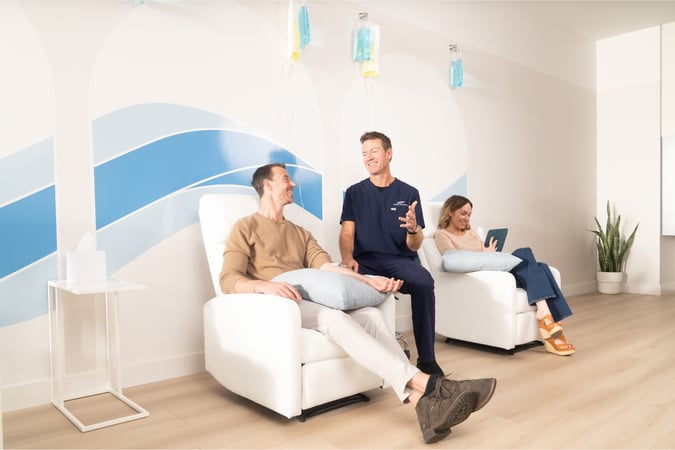 Dr. Florie and a patient relax in white recliner chairs receiving IV therapy while a clinician in scrubs sits between them, smiling and chatting