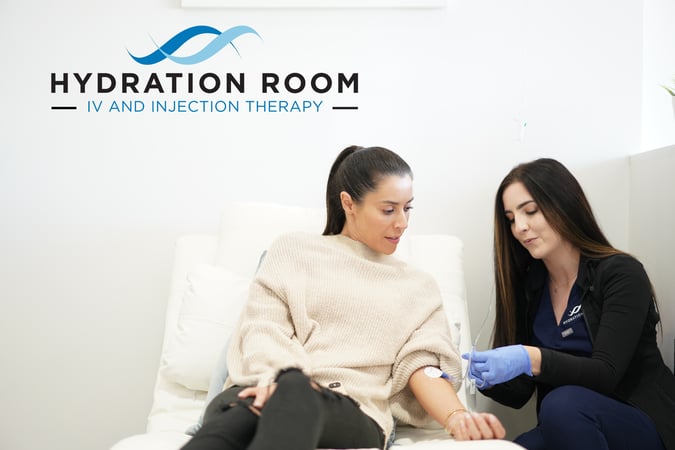 A woman reclines in a white chair while a clinician wearing gloves inserts an IV line in her arm.