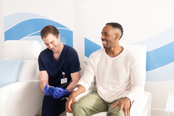 A smiling healthcare worker in navy scrubs and blue gloves prepares an injection for a seated patient in a bright, modern clinic room